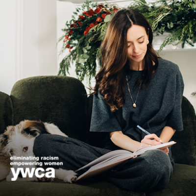 Woman sitting down on the couch with her dog next to her writing down her goals in a journal